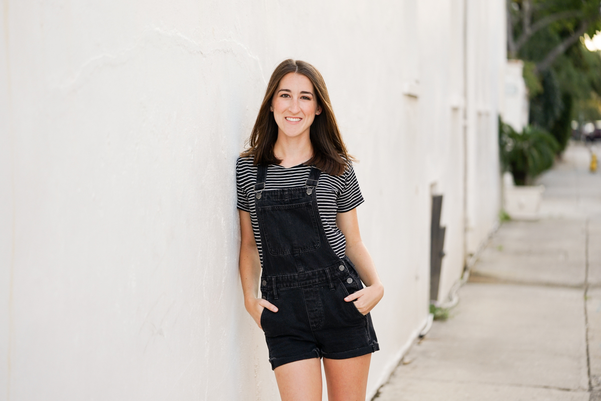 Girl in black overalls leaning against a white wall