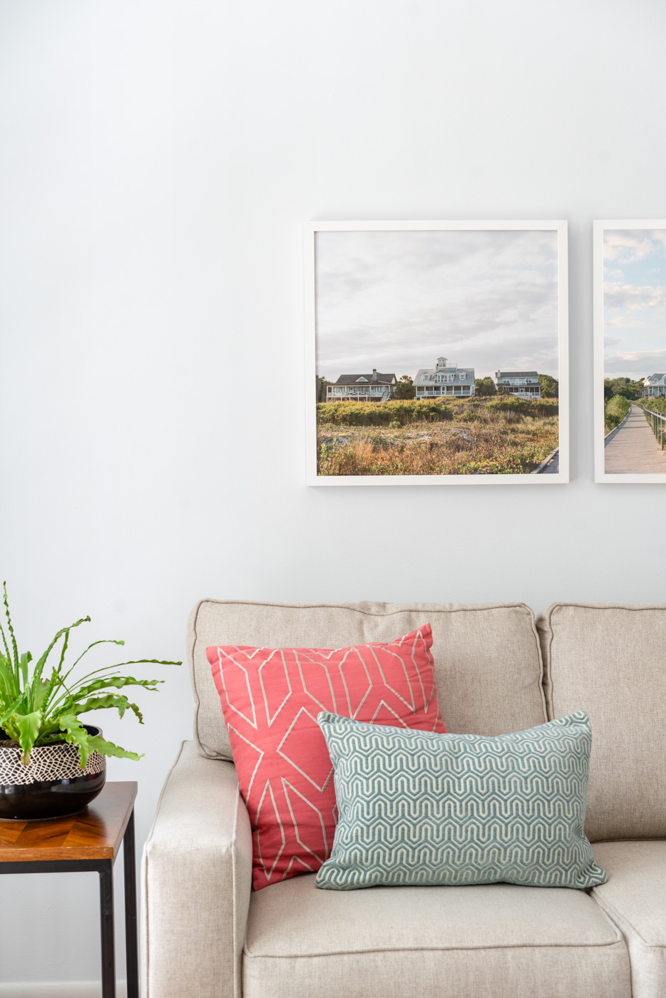 A diptych photograph of beach homes and a walkway on Sullivan's Island, SC hanging over a beige couch with 2 colorful pillows and a plant on the side table