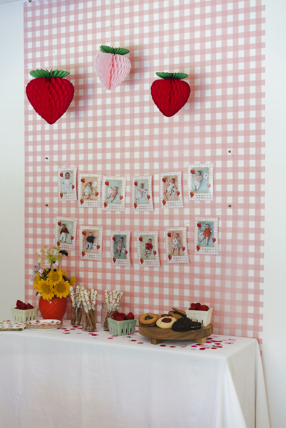 Pink and white gingham backdrop with hanging paper strawberries above. On the backdrop is a string of baby photos, one for each month of the baby's first year. On the table below is a vase of flowers, plate of cookies, and jars of pretzels. 
