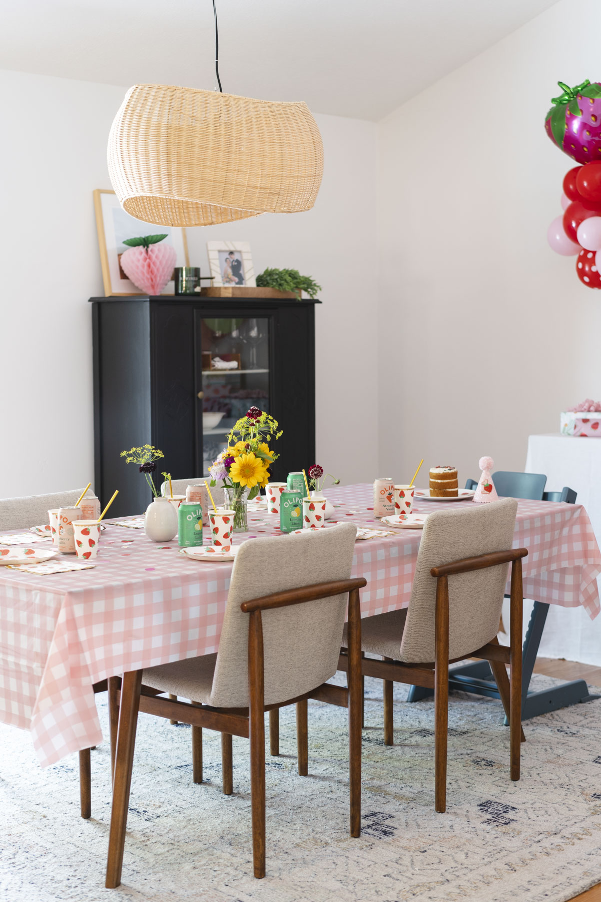 Set up of the birthday table with a pink and white gingham table cloth, vase of sunflowers, and strawberry patterned plates and cups. 