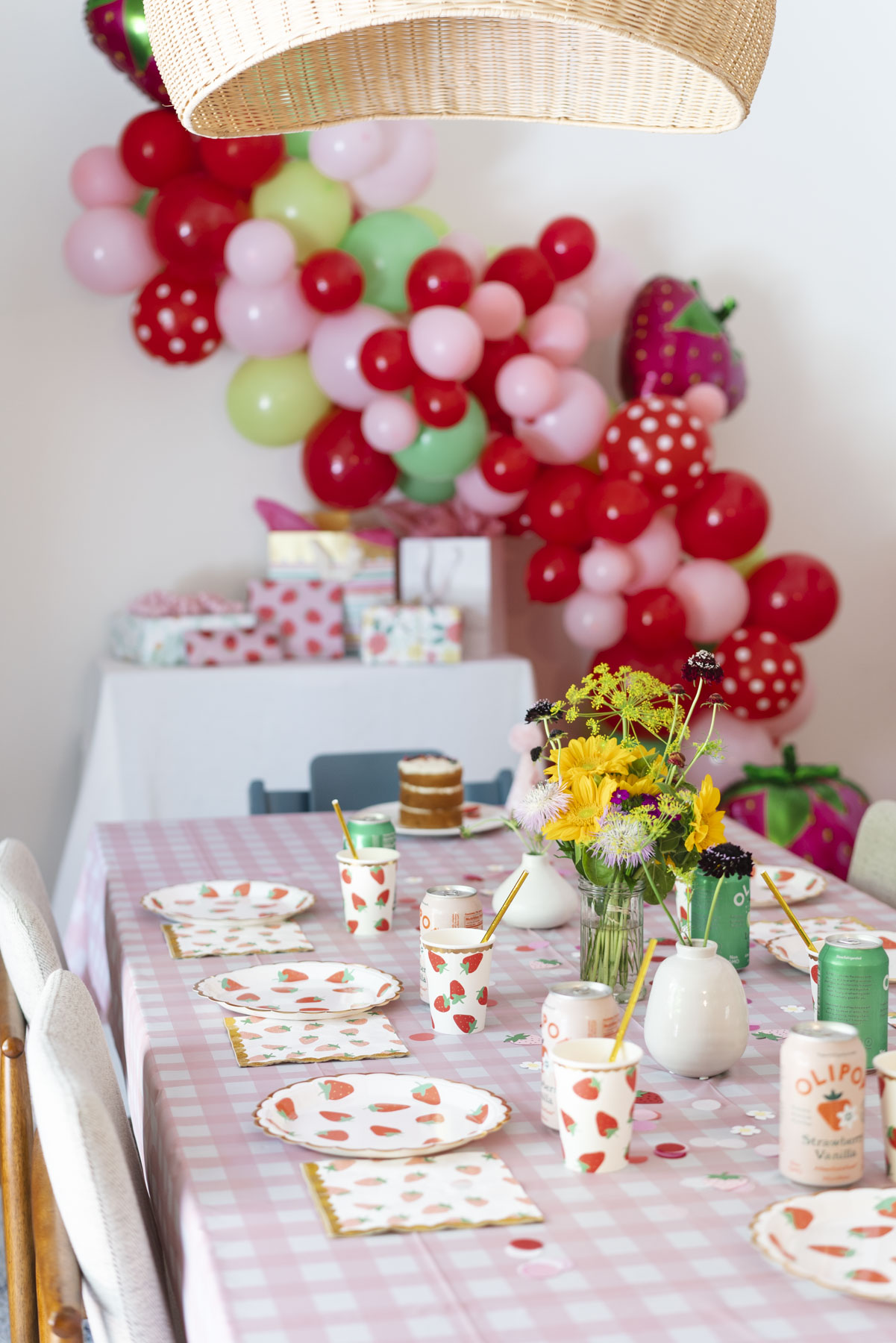 Focus of the birthday table with a vase of yellow flowers, and strawberry patterned cups and plates. Behind is the strawberry balloon arch. 
