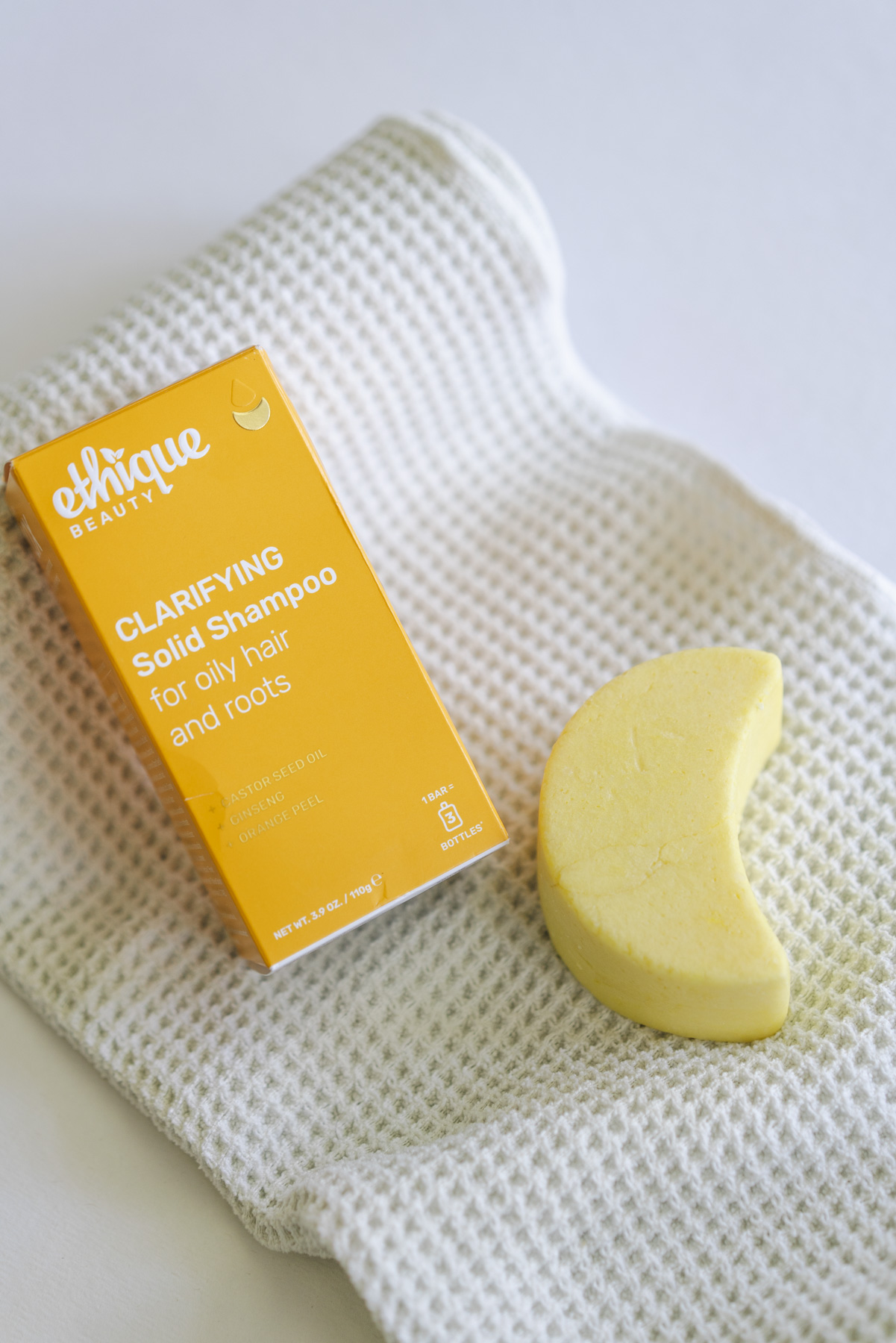 Flat lay photo of a yellow shampoo bar sitting next to the box it came in. The shampoo bar is yellow and shaped like a crescent moon. The bar and box are laid on top of a white, waffle textured towel. 