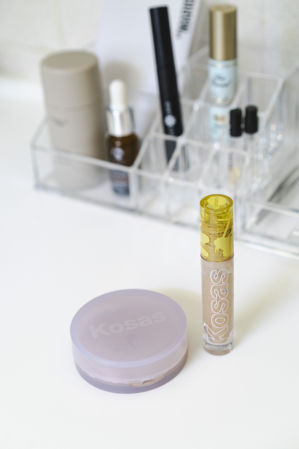 Close up photo of Kosas makeup sitting on a clean, white countertop. In the background in a clear, plastic makeup organizer that is filled with a few other products. 