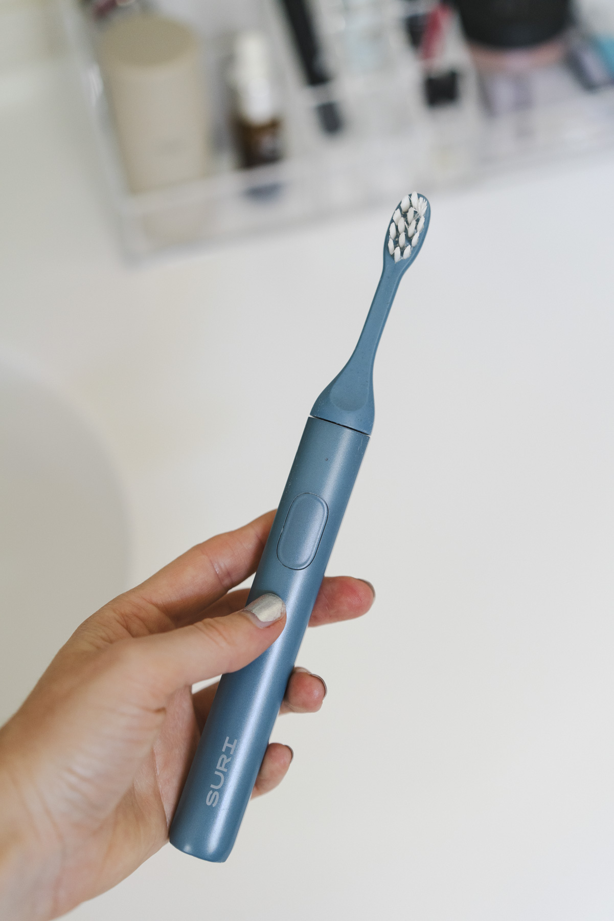 Close up photo of a hand holding the SURI toothbrush. The toothbrush is blue with white bristles. 