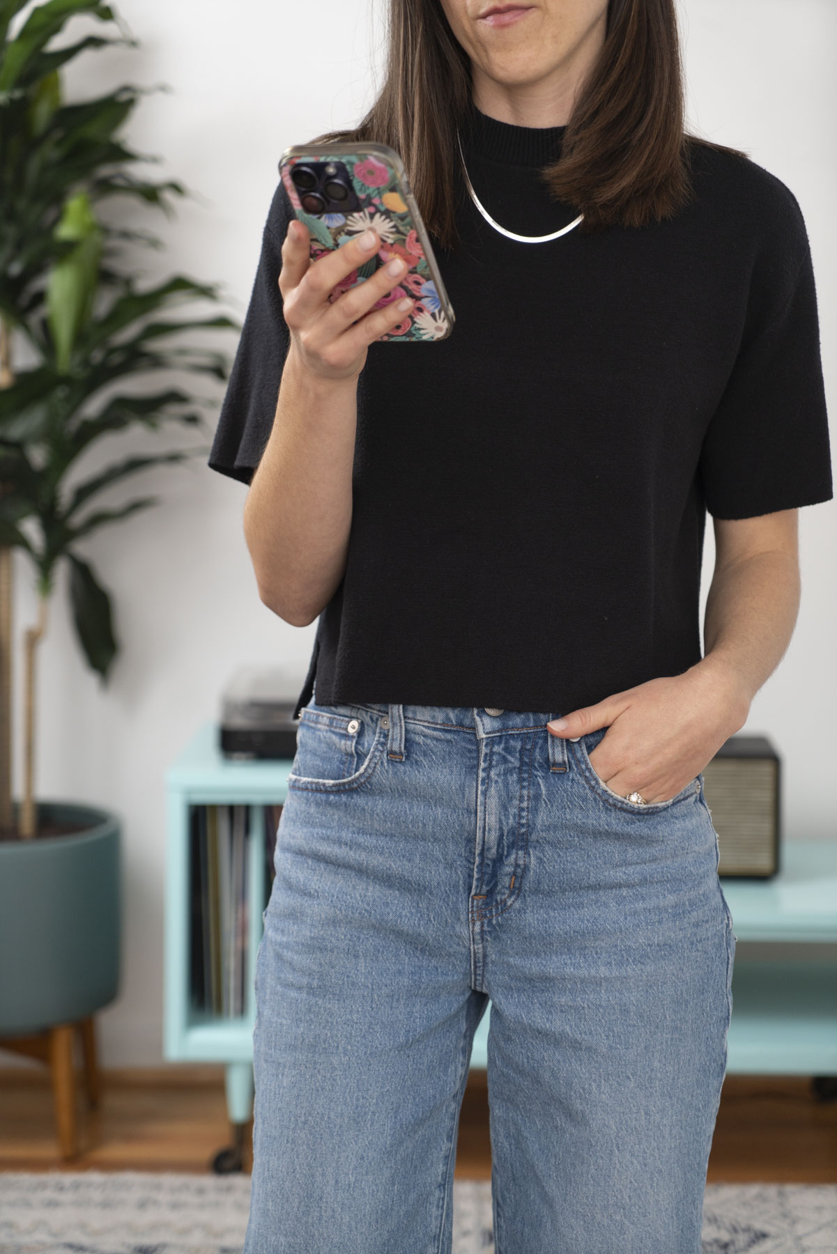 View of Abby from the neck down, she is holding her phone and wearing a black short sleeve top, jeans, and a silver necklace.