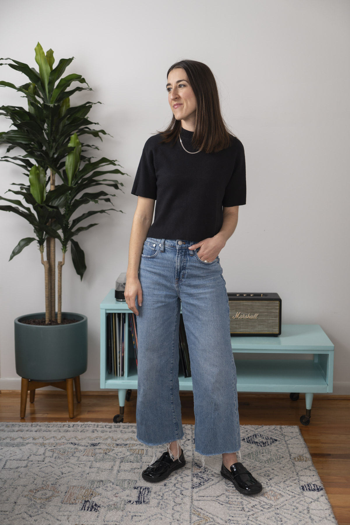 Abby posing in front of a large potted plant and record player cabinet. She is wearing a black short sleeve top and wide leg jeans.