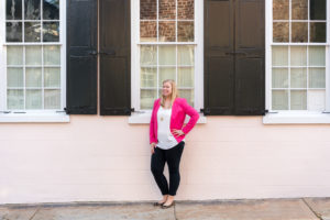 Woman in a pink blazer and black pants standing against a pink house with black shutters