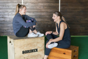 A trainer and a client sitting on wood blocks in a gym laughing