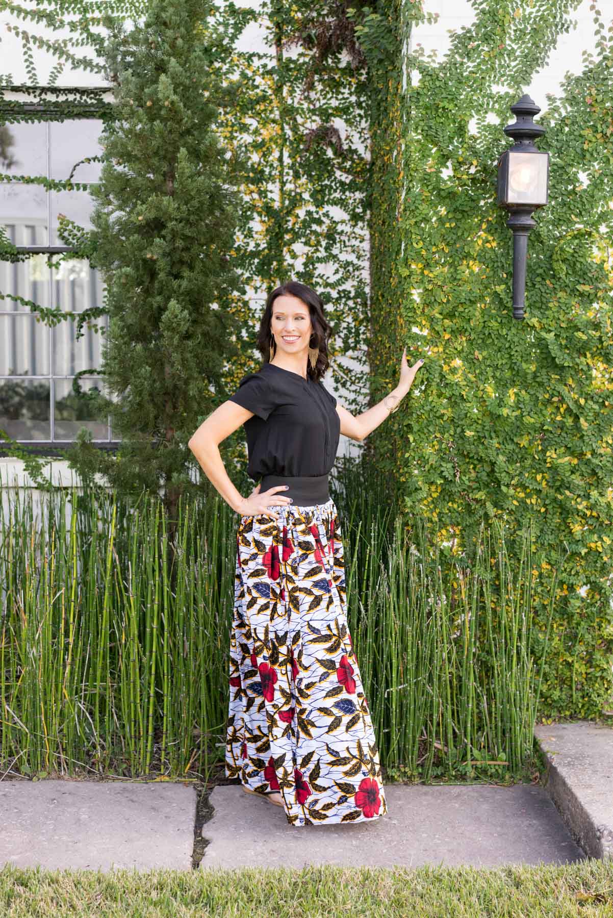 Full-length portrait of a woman wearing a bold, patterned skirt and black top leaning against a wall covered in ivy. An example in a post about how to look confident in your brand photos. 