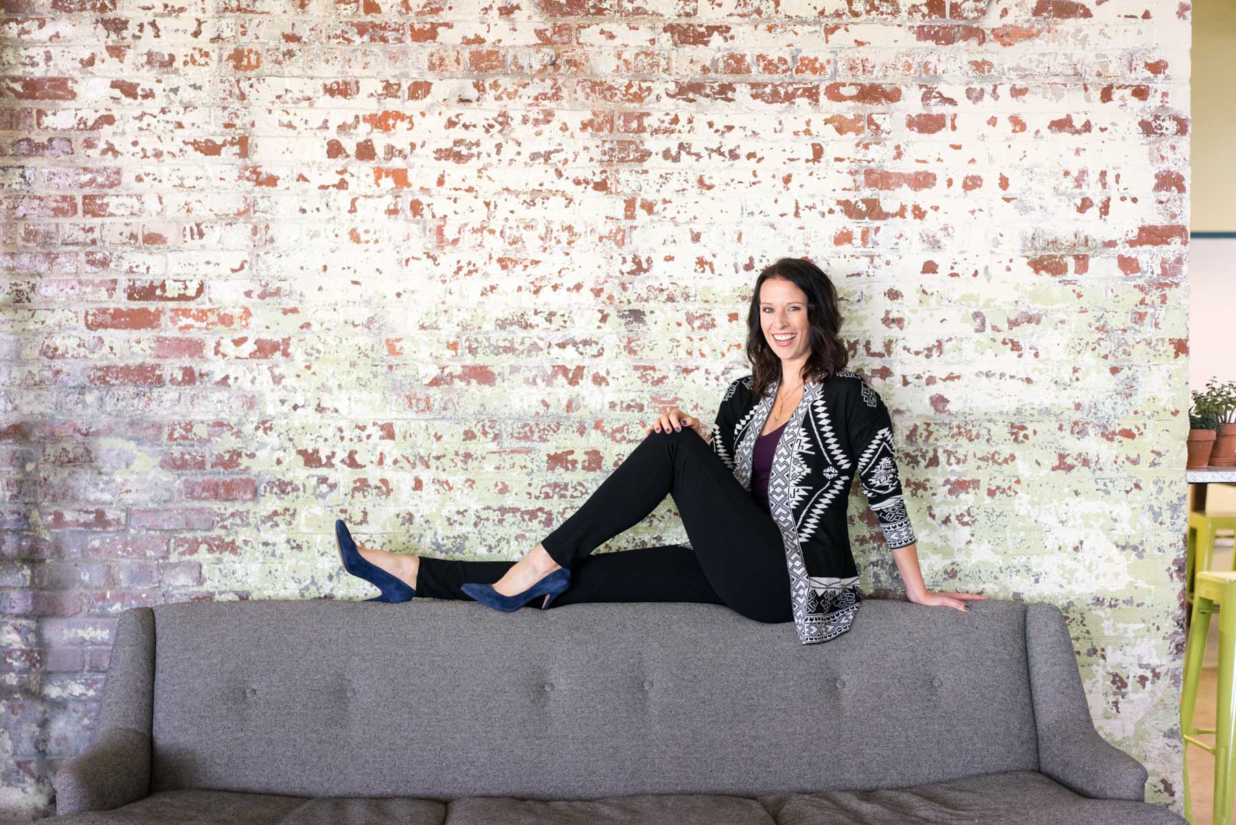 A woman displaying how to look confident in brand photos. She's posed atop a couch against a distressed brick wall wearing blue velvet heels and a black outfit. 
