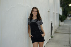 Girl in black overalls leaning against a white wall