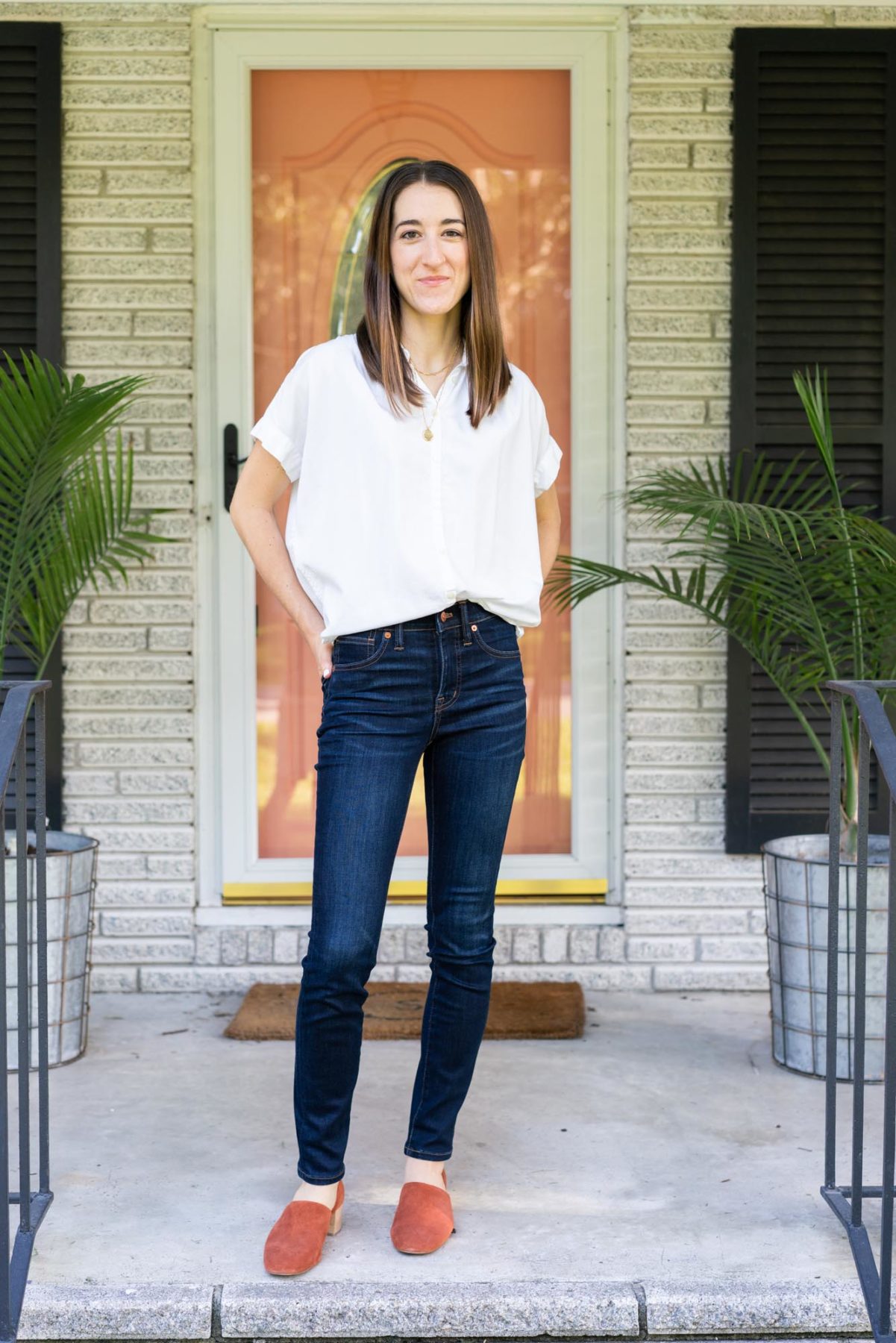 Woman standing on a front porch wearing an outfit from her Madewell capsule collection, including a white Madewell Central Shirt, 10" high-rise dark denim, and suede mules