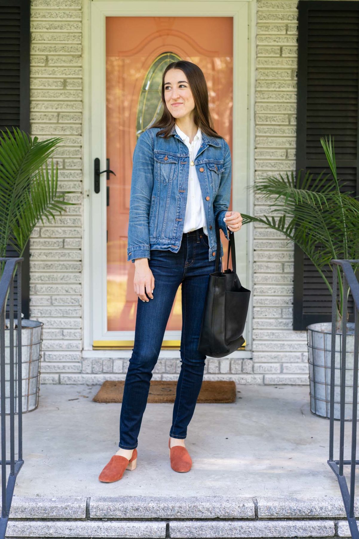 Woman standing on her front porch wearing Madewell denim, a denim jacket, button up shirt, and Madewell tote bag