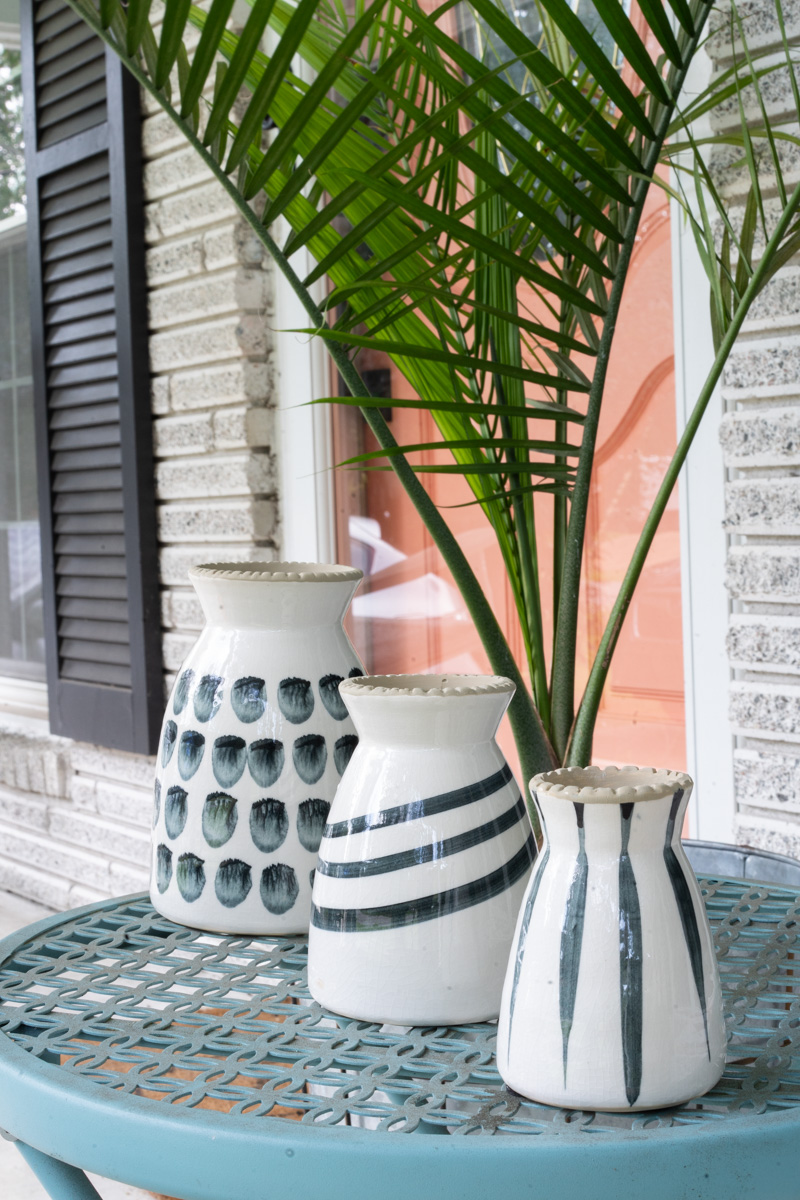 Three black and white patterned vases on a blue outdoor table with a palm plant and a pink door in the background. The purpose is to show how aperture in photography works. 