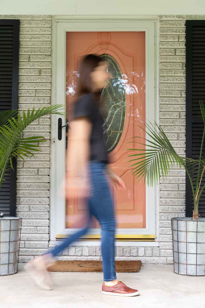 A woman walking across a front porch with a pink door with her motion blurred. The purpose is to show how shutter speed in photography works. 