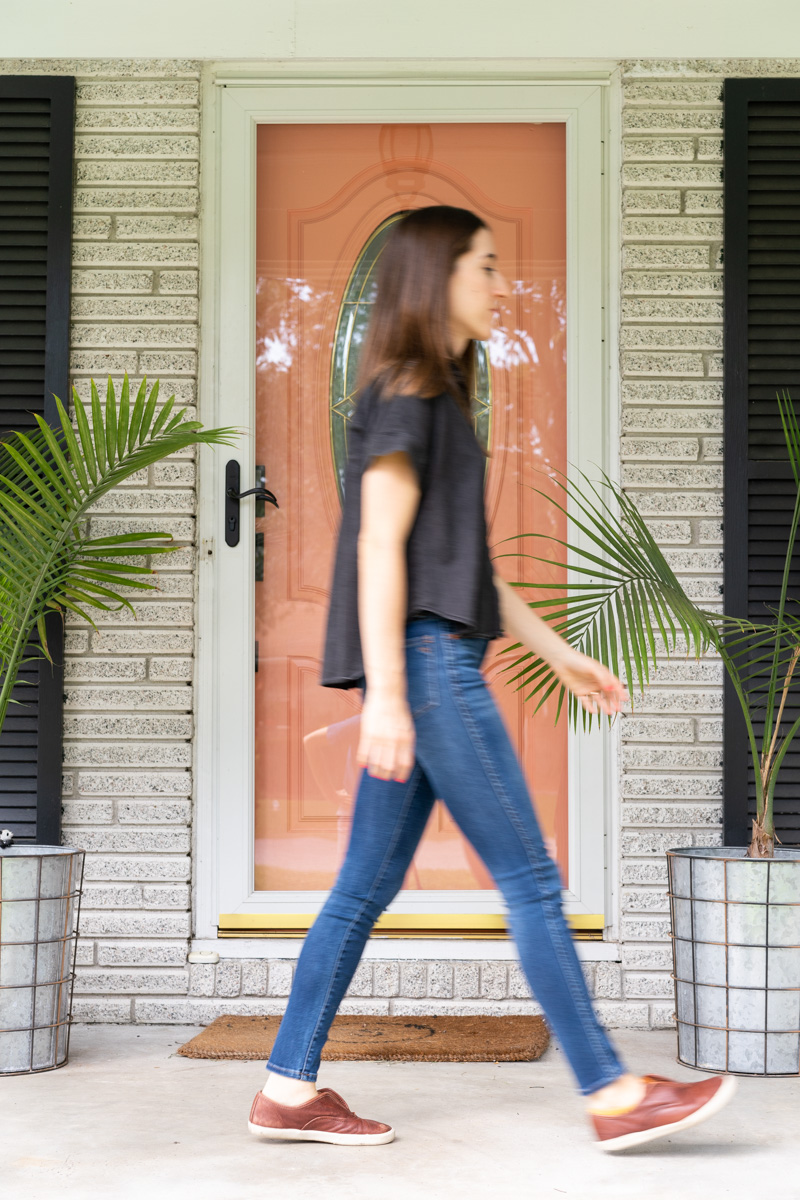 A woman walking across a front porch with a pink door with her motion blurred. The purpose is to show how shutter speed in photography works. 