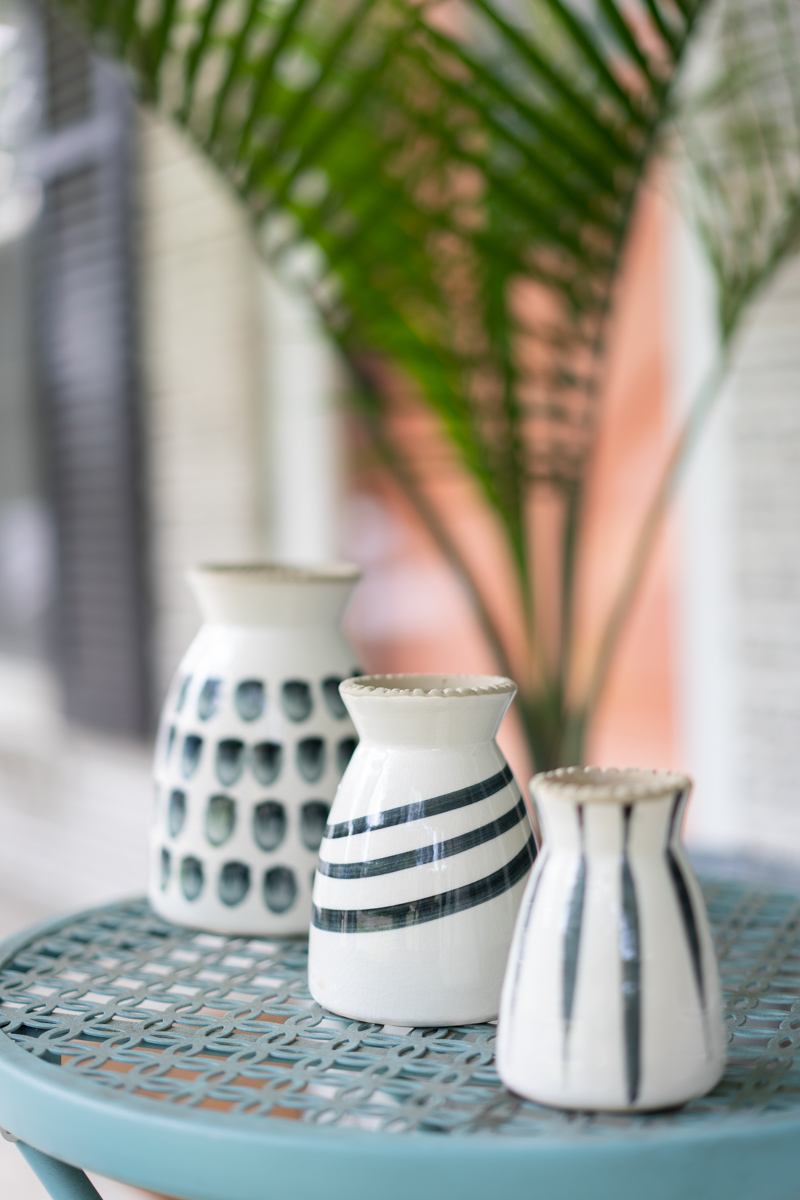Three black and white patterned vases on a blue outdoor table with a palm plant and a pink door in the background. The purpose is to show how aperture in photography works. 