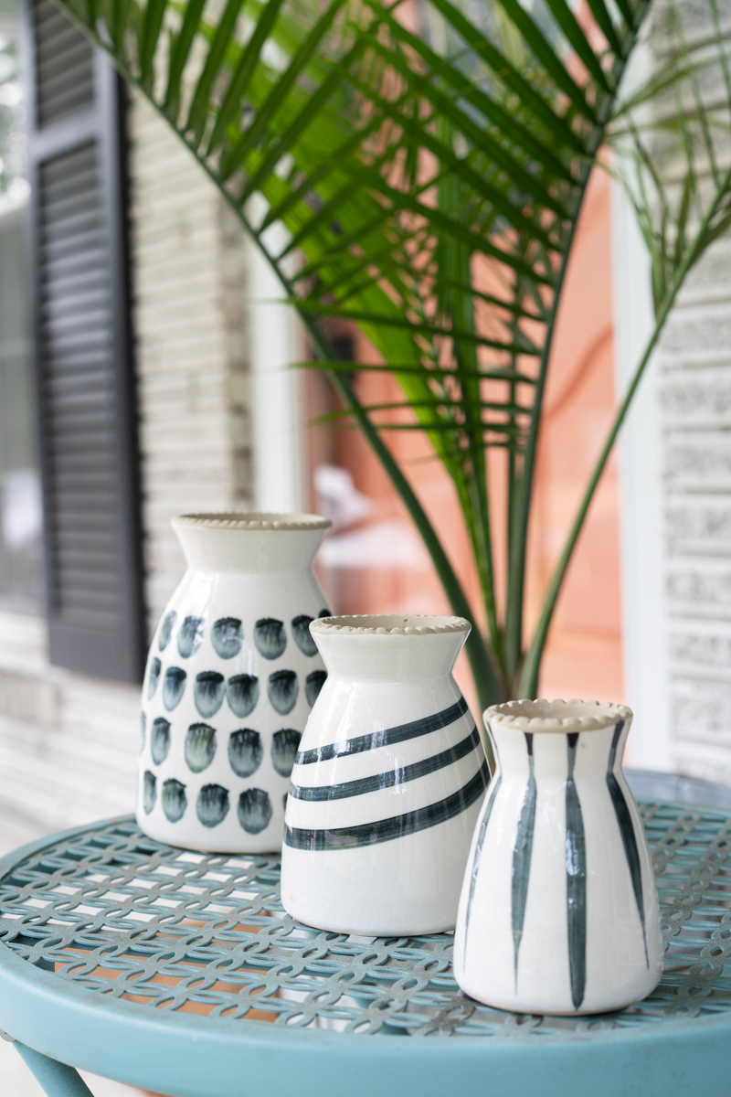 Three black and white patterned vases on a blue outdoor table with a palm plant and a pink door in the background. The purpose is to show how aperture in photography works. 