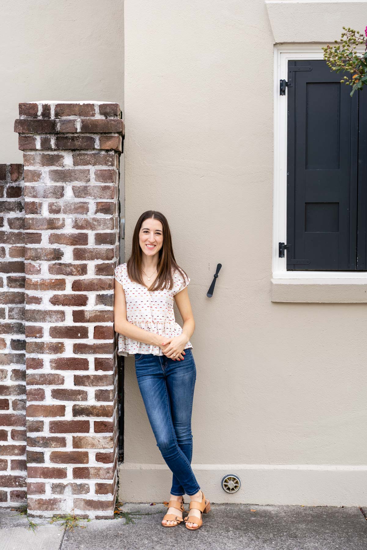 Woman in denim jeans and a white blouse demonstrating how to pose for a photoshoot by crossing her ankles and lightly leaning against a brick wall