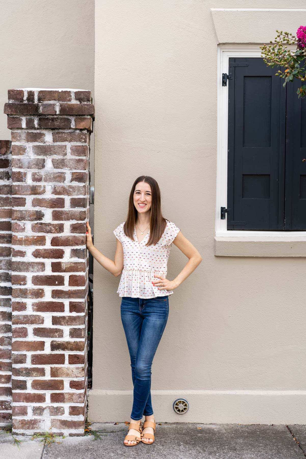 Woman in denim jeans and a white blouse demonstrating how to pose for a photoshoot by crossing her ankles and lightly placing a hand on a nearby brick wall