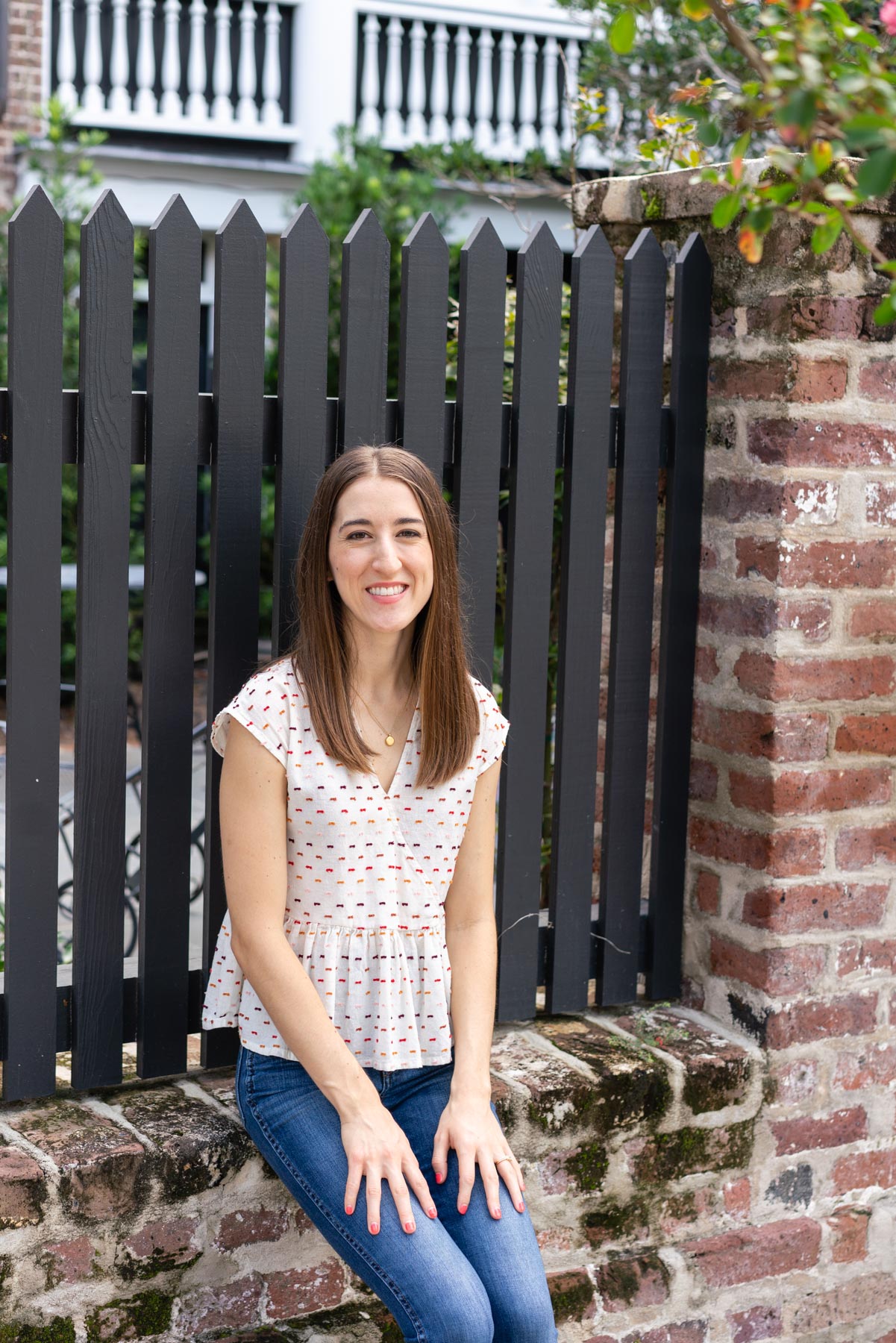 Woman in denim jeans and a white blouse sitting on the edge of a brick wall with her arms stiffly at her side demonstrating how not to pose for a photoshoot 