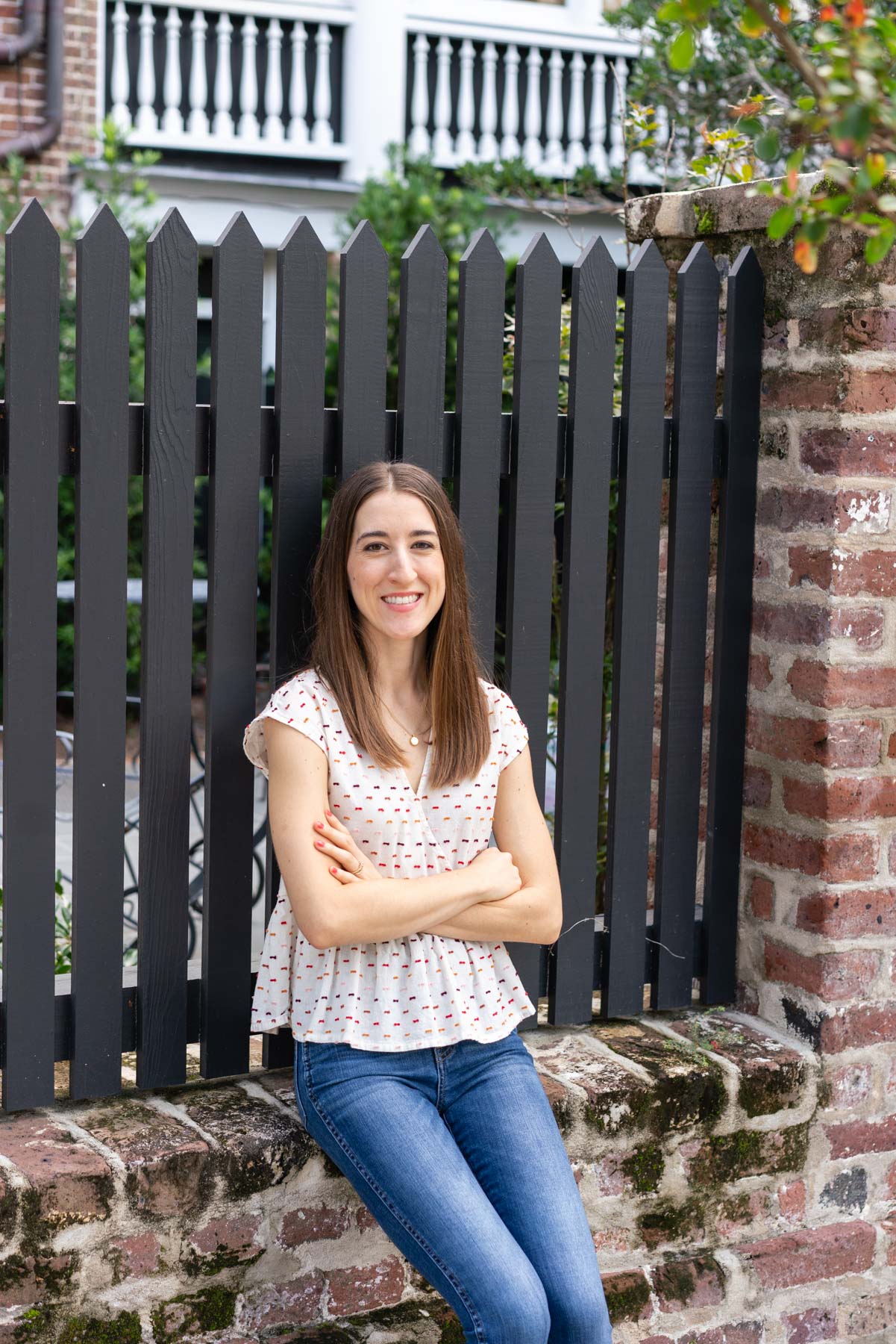 Woman in denim jeans and a white blouse sitting on the edge of a brick wall with her arms crossed demonstrating how not to pose for a photoshoot 