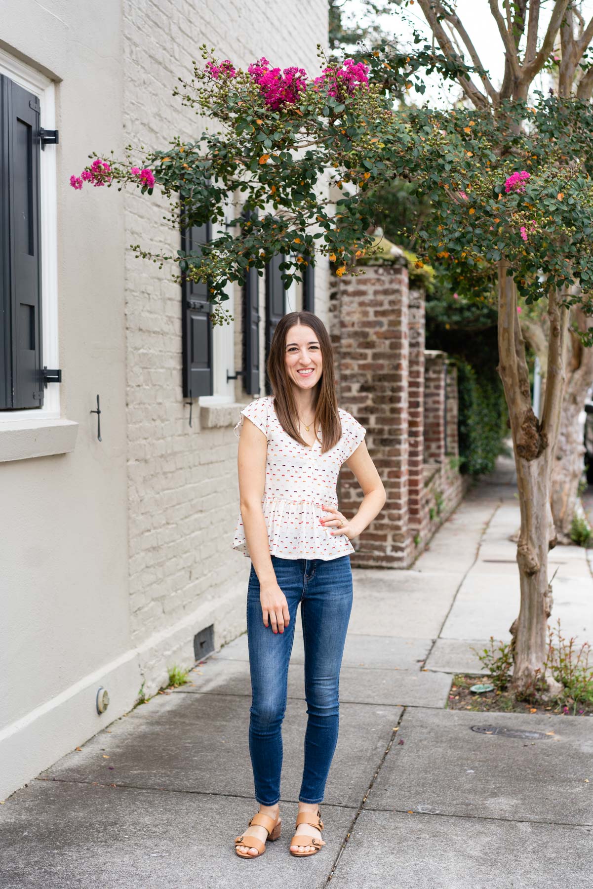 Woman in denim jeans and a white blouse demonstrating how to pose for a photoshoot by placing one hand on her hip and smiling naturally at the camera
