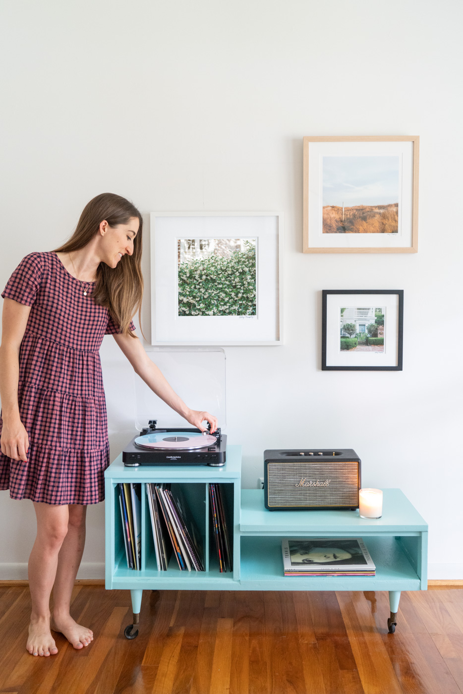 Three framed photographs of scenes in Charleston, South Carolina hanging above a mid-century modern record player table. A woman stands to the left side putting a record on the record player