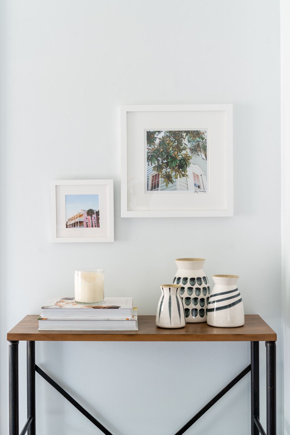 Two photographs in white frames hanging on the wall over a bookcase styled with books, a candle, and 3 black and white vases. 
