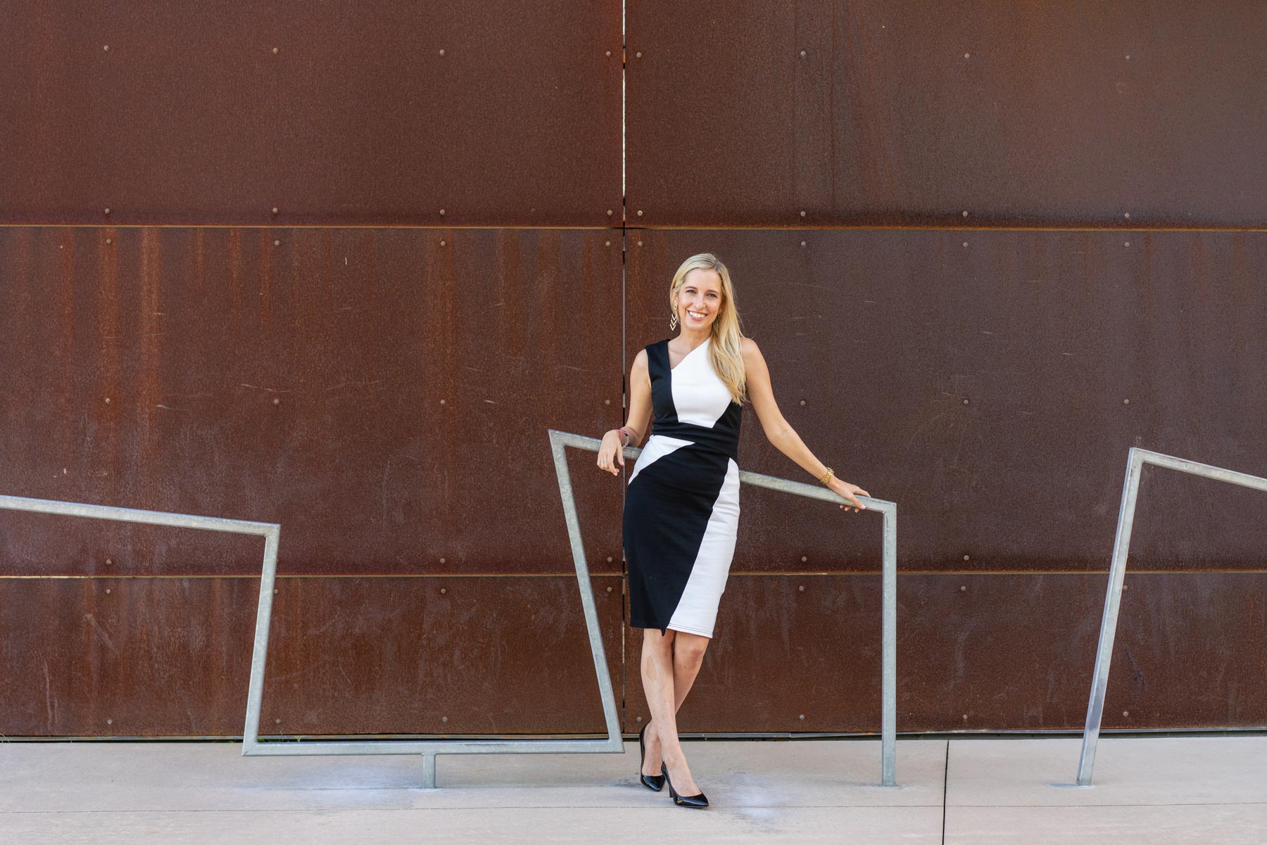 Marketing brand headshot of a woman in a black and white dress standing in front of a rust-colored, industrial wall 