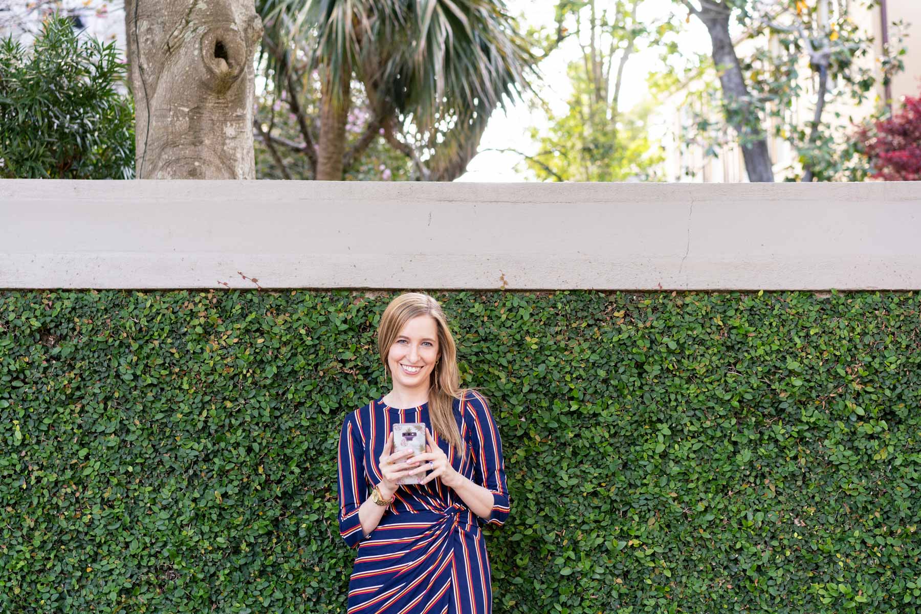 Woman in a striped dress standing against an ivy wall holding her phone and smiling