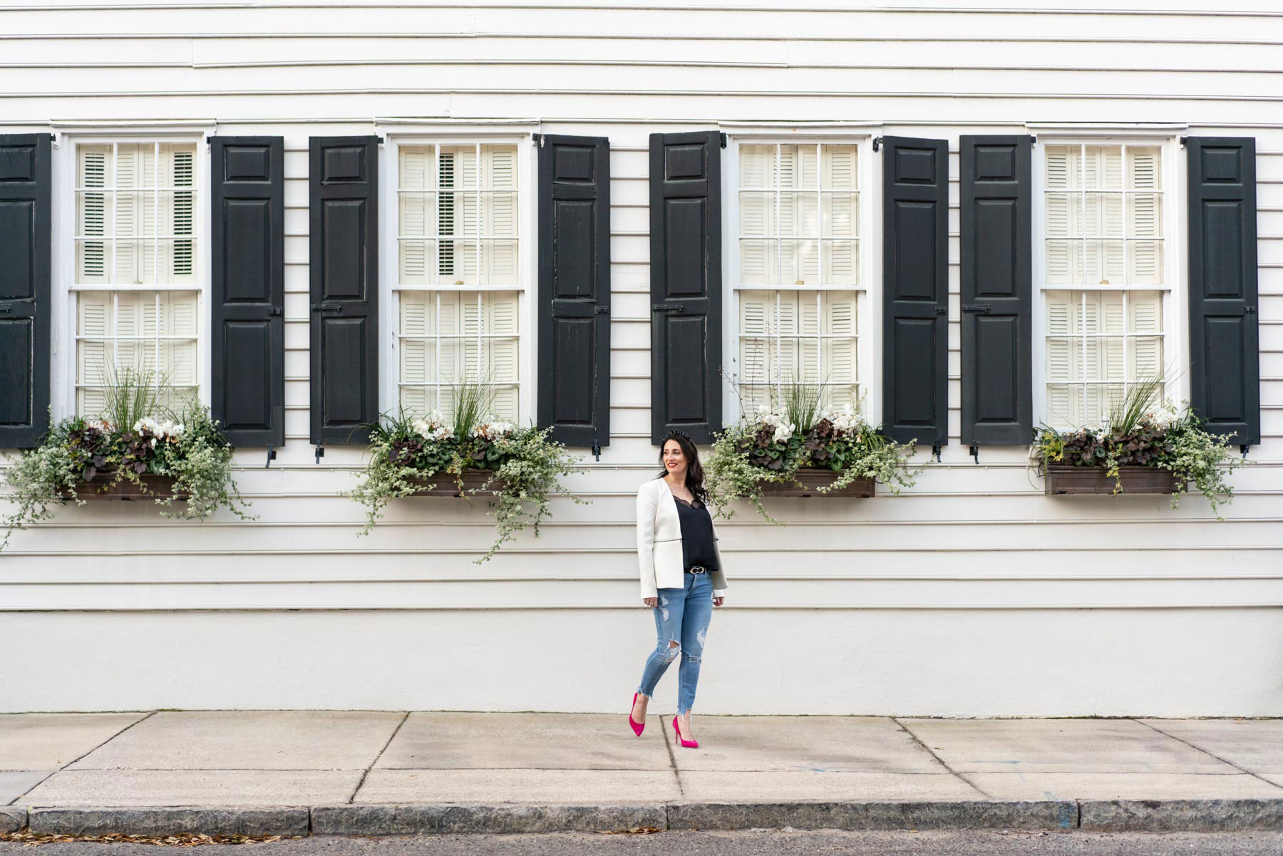 Woman in a white blazer, hot pink heels, and ripped jeans standing in front of a white wall with black shutters and window boxes in Charleston, South Carolina. Brand photography by Abby Murphy.