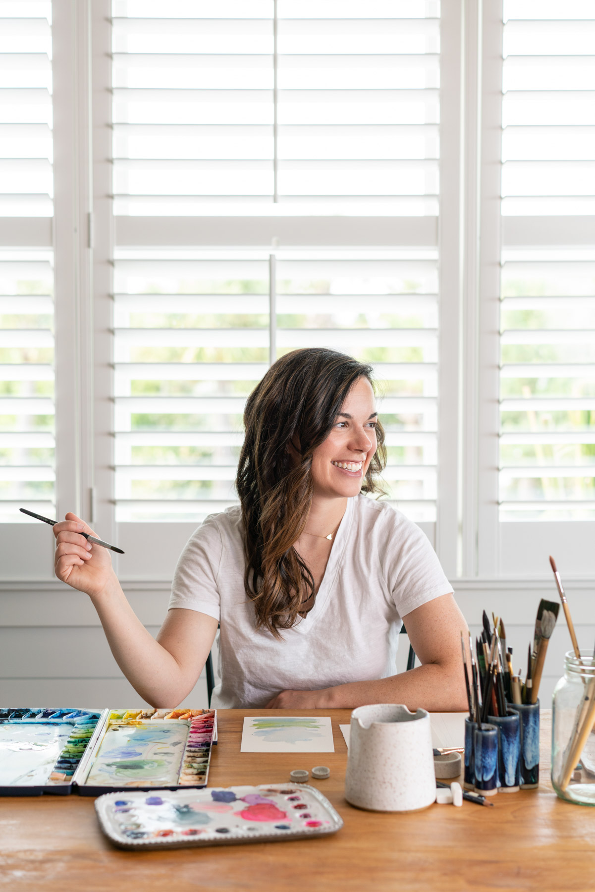 Brand photo of a female artist sitting at a table smiling with watercolor painting supplies