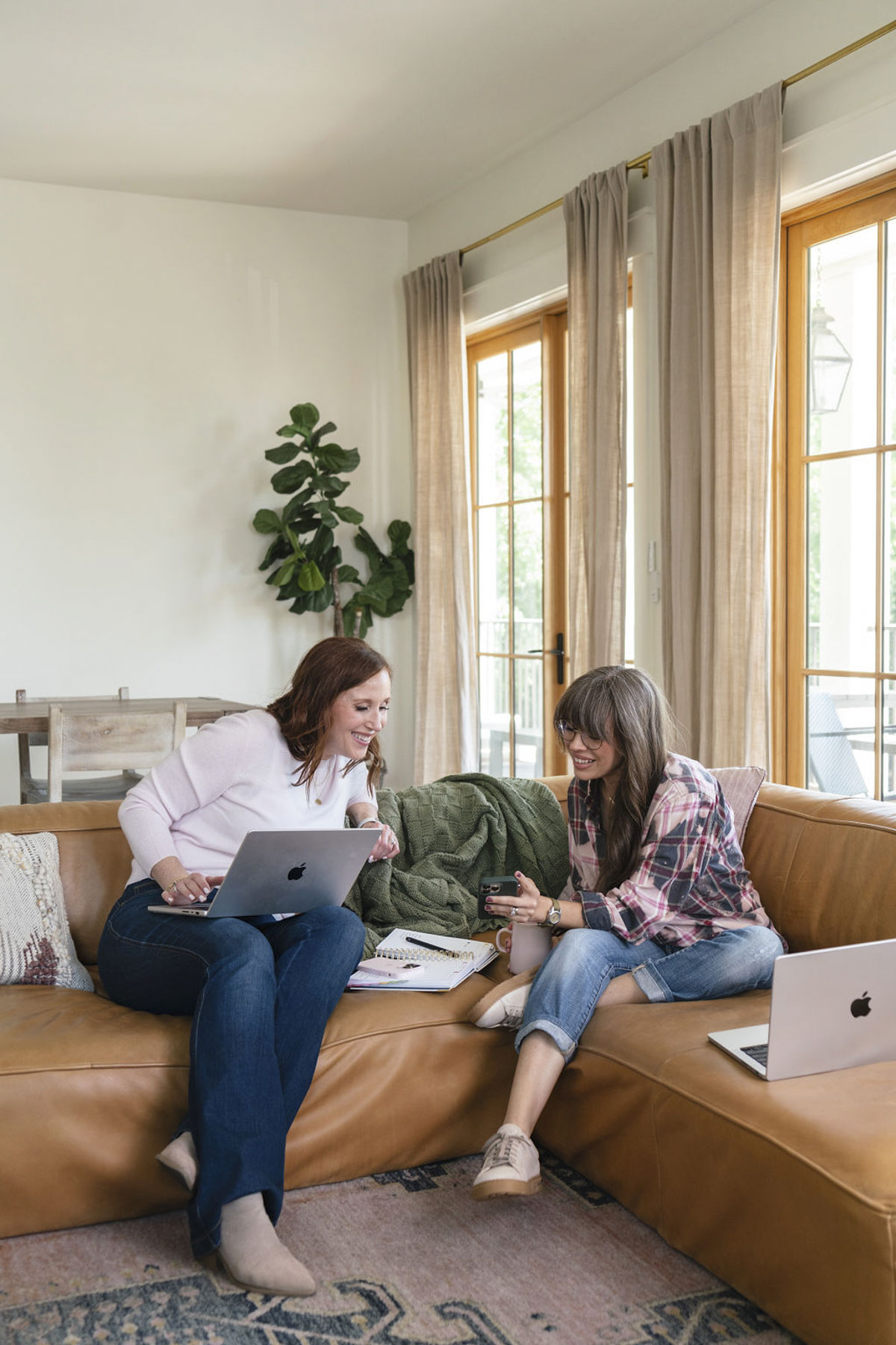 Two women sitting on the sofa and smiling. One has a laptop in her lap. The other is holding her phone, and they are both looking at the phone. 